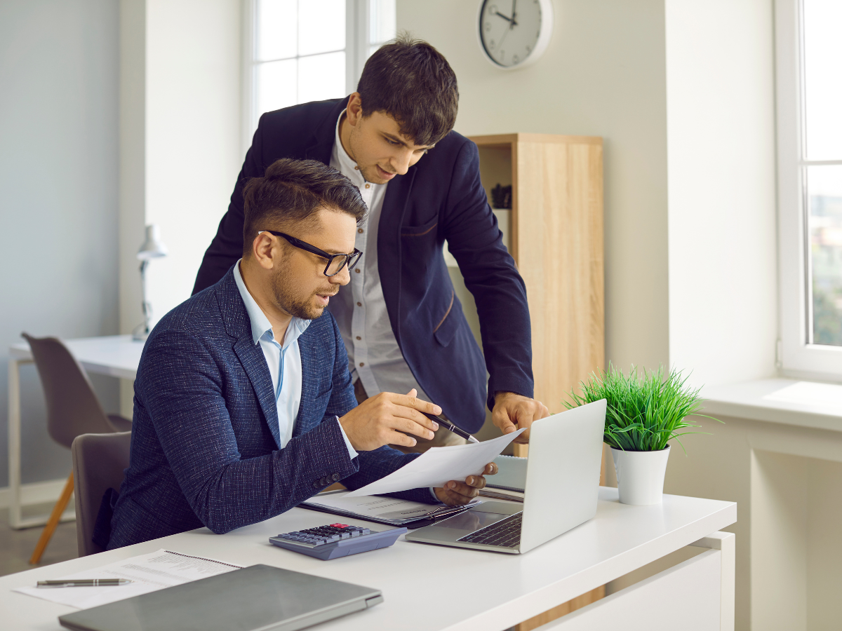 Two men in business attire review documents together at a white desk with a laptop and calculator present in a bright office.